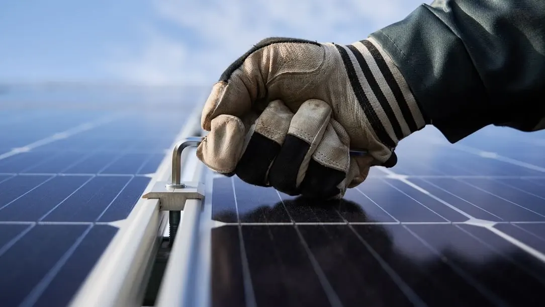 Nahaufnahme einer Hand mit Arbeitshandschuh am Aluminiumrahmen eines Solarmoduls